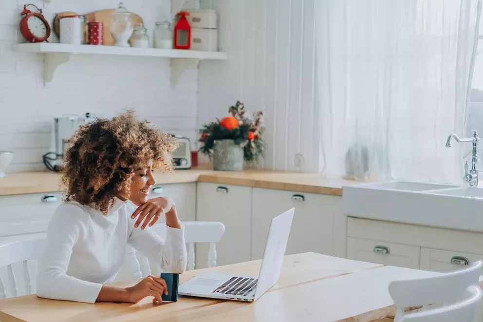 smiling woman sitting at kitchen table with laptop and credit card smiling woman sitting at kitchen table with laptop and credit card