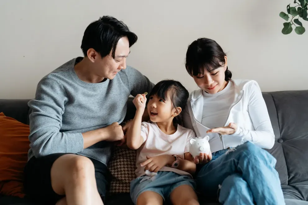 Happy family of three sitting together on gray couch, parents with piggy bank and young child between them Happy family of three sitting together on gray couch, parents with piggy bank and young child between them