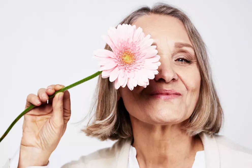 smiling senior woman holding pink daisy over one eye smiling senior woman holding pink daisy over one eye