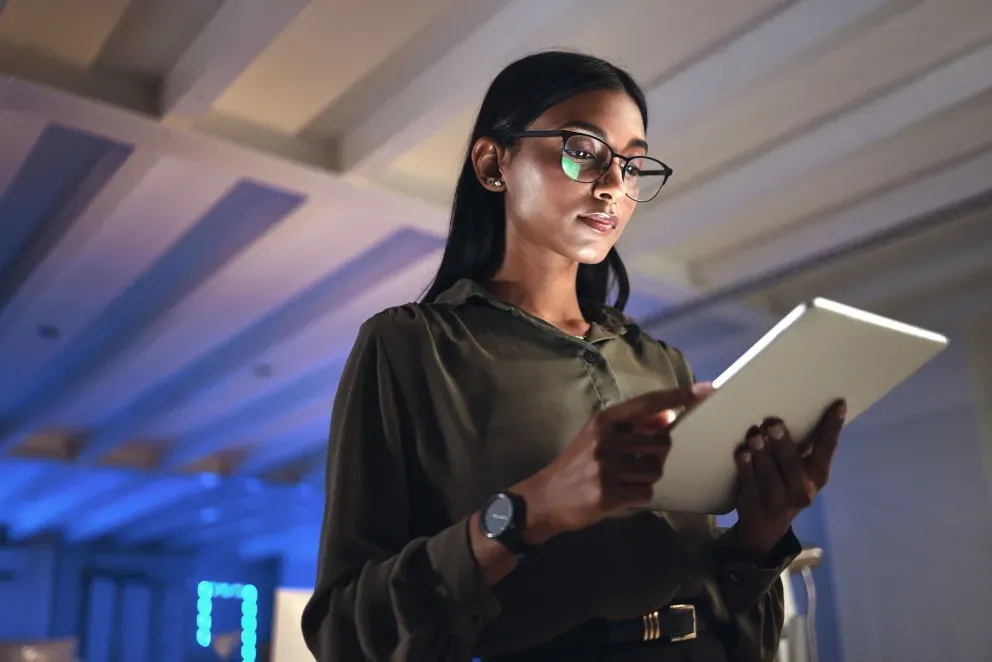 Young woman with glasses holding a tablet, looking up in a dimly lit modern office setting. Young woman with glasses holding a tablet, looking up in a dimly lit modern office setting.