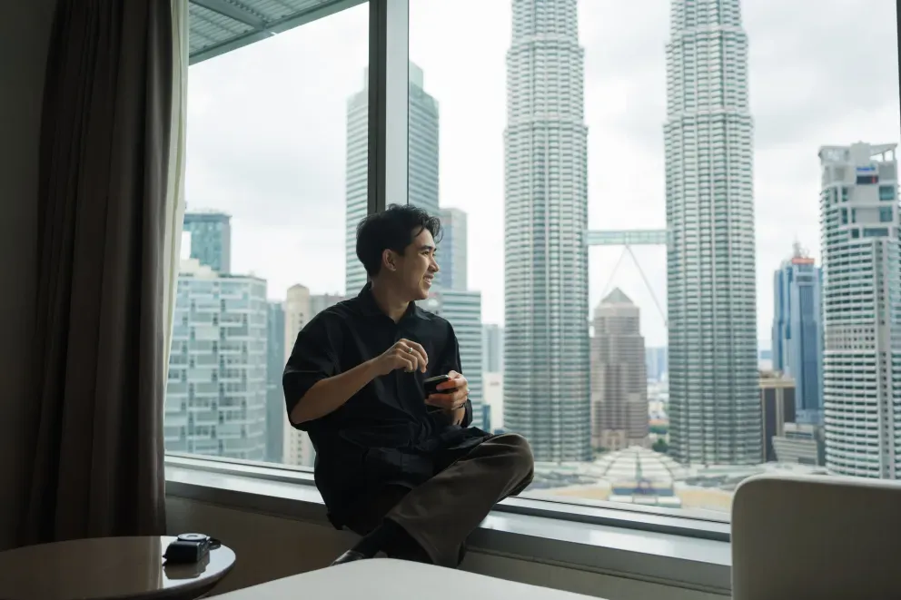 Man sitting on a windowsill holding a cup, with Kuala Lumpur's Petronas Towers visible behind him. Man sitting on a windowsill holding a cup, with Kuala Lumpur's Petronas Towers visible behind him.