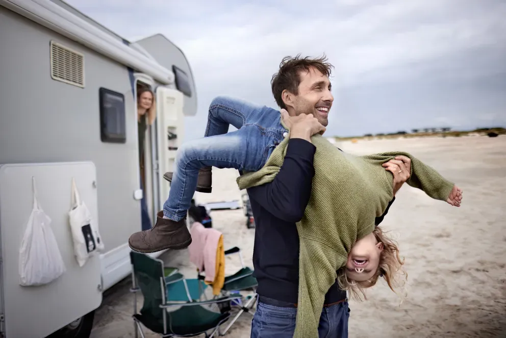 Man carrying a child, laughing beside a parked RV on a sandy beach. Man carrying a child, laughing beside a parked RV on a sandy beach.