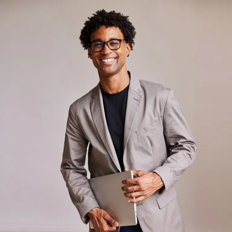 Young man with curly hair and glasses wearing a gray blazer, holding a laptop, smiling against a neutral background. Young man with curly hair and glasses wearing a gray blazer, holding a laptop, smiling against a neutral background.