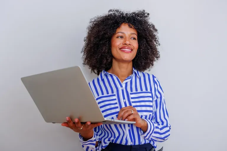Smiling woman with curly hair in blue striped shirt holding laptop against gray background Smiling woman with curly hair in blue striped shirt holding laptop against gray background