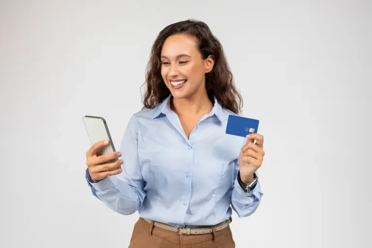 Smiling young woman holding a smartphone and blue credit card against a light gray background. Smiling young woman holding a smartphone and blue credit card against a light gray background.