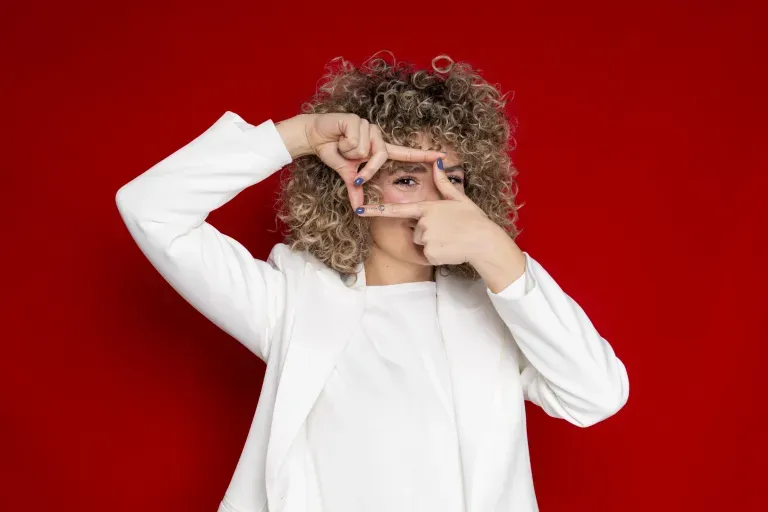 Person with curly blonde hair in a white outfit framing one eye with their fingers against a red background. Person with curly blonde hair in a white outfit framing one eye with their fingers against a red background.