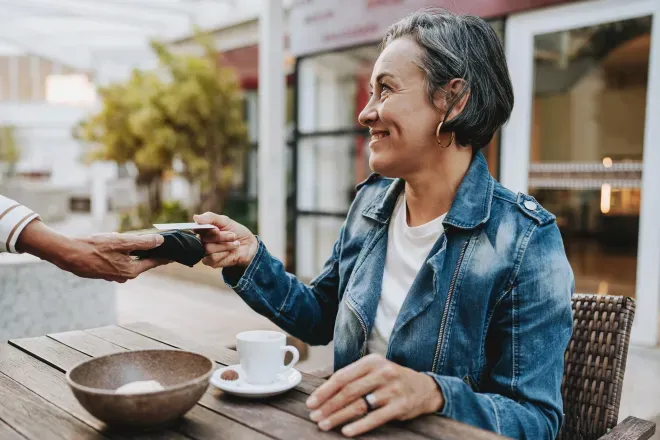 Smiling older woman tapping her phone to a card reader at an outdoor café table. Smiling older woman tapping her phone to a card reader at an outdoor café table.