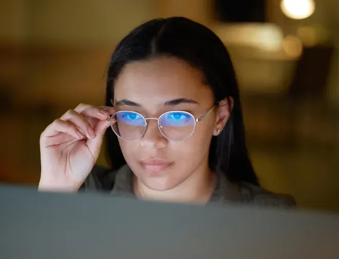 Young woman peering over glasses at a computer screen in a dimly lit office. Young woman peering over glasses at a computer screen in a dimly lit office.