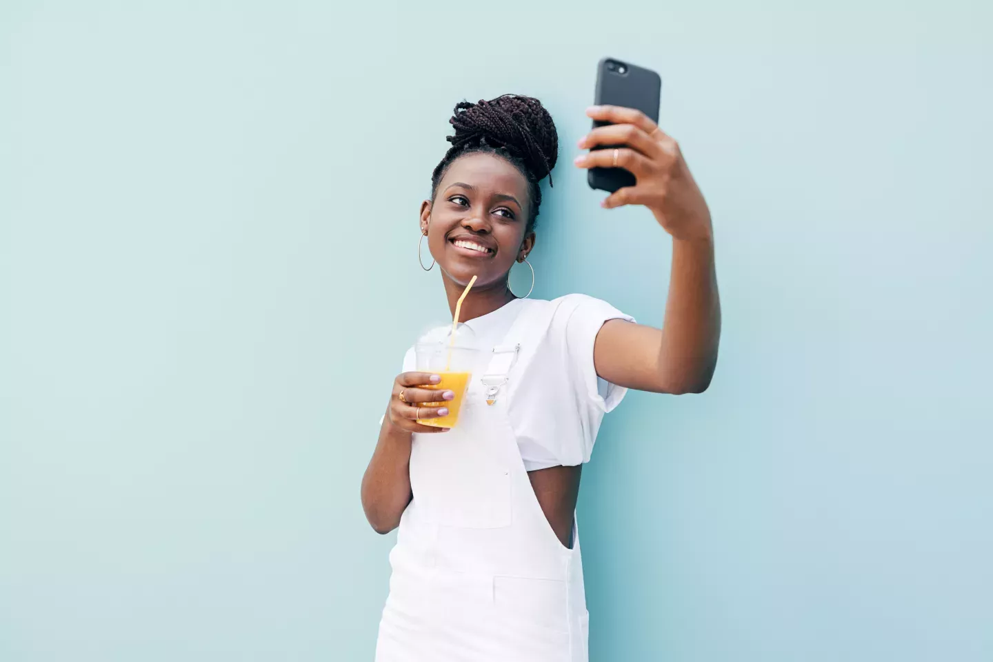 smiling young woman leaning on light blue wall holding orange juice holding cell phone smiling young woman leaning on light blue wall holding orange juice holding cell phone