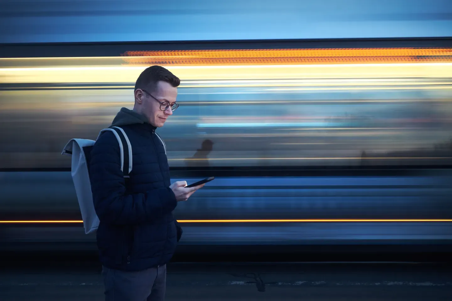 Man in dark jacket with phone as blue train speeds past in motion blur Man in dark jacket with phone as blue train speeds past in motion blur