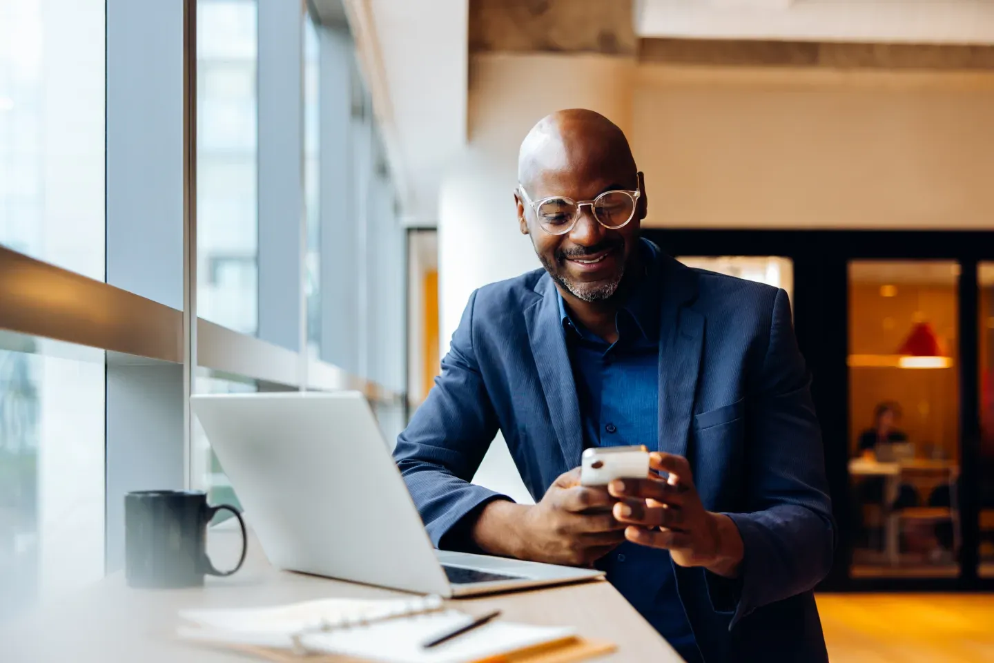 Smiling businessman in a blue suit holding a phone at a bright office desk with a laptop. Smiling businessman in a blue suit holding a phone at a bright office desk with a laptop.