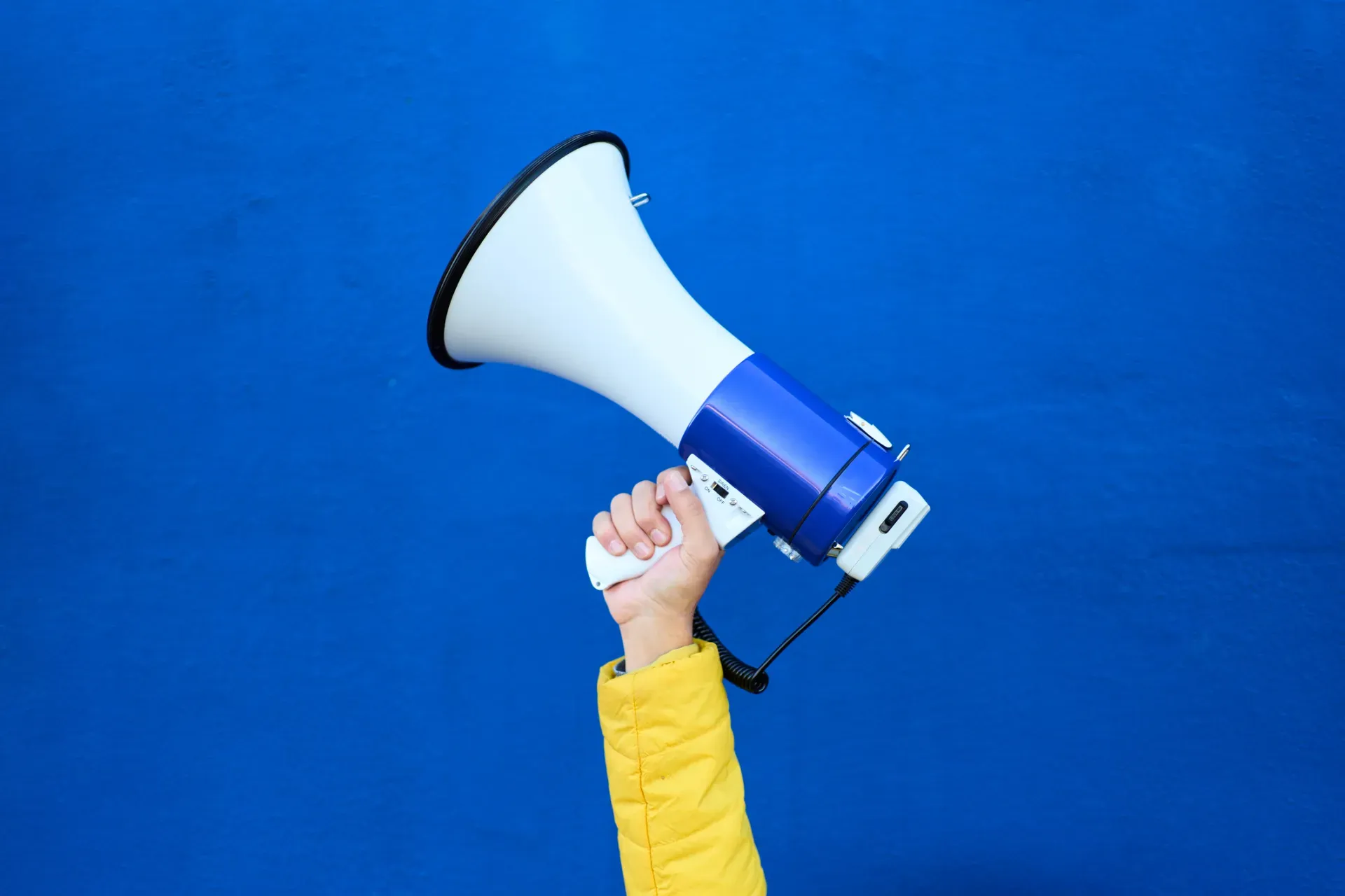 Hand holding white and blue megaphone against bright blue background Hand holding white and blue megaphone against bright blue background