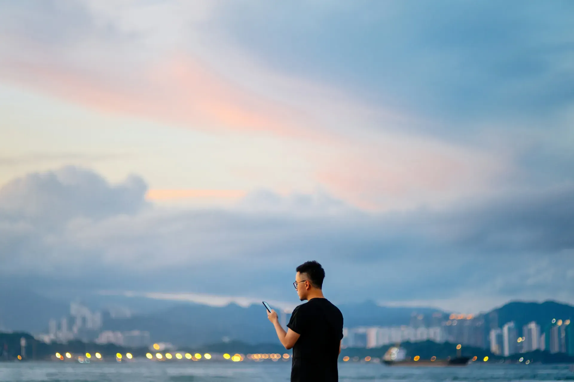 Person with phone silhouetted against twilight sky overlooking illuminated waterfront cityscape Person with phone silhouetted against twilight sky overlooking illuminated waterfront cityscape
