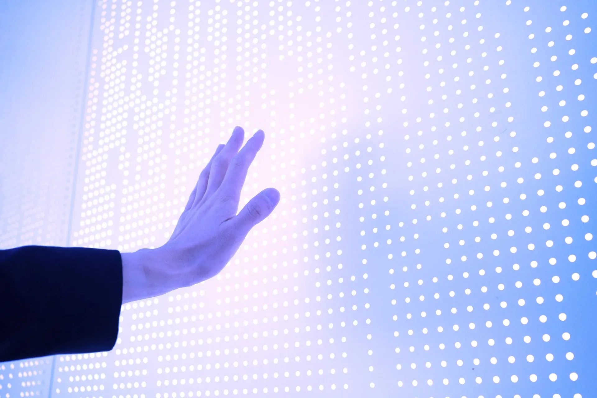 Hand reaching toward a glowing white LED dot-matrix panel with blue and purple light. Hand reaching toward a glowing white LED dot-matrix panel with blue and purple light.