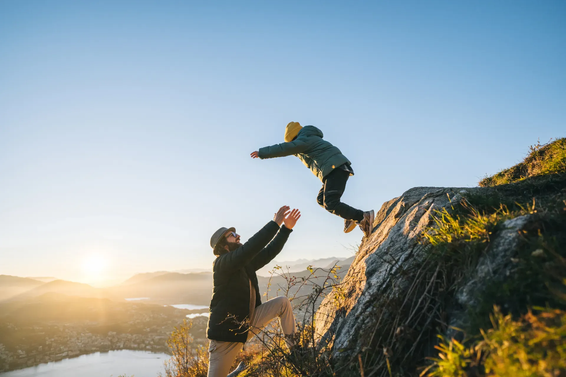 Adult lifting a child jumping off a rocky cliff at sunset, mountains in the background. Adult lifting a child jumping off a rocky cliff at sunset, mountains in the background.
